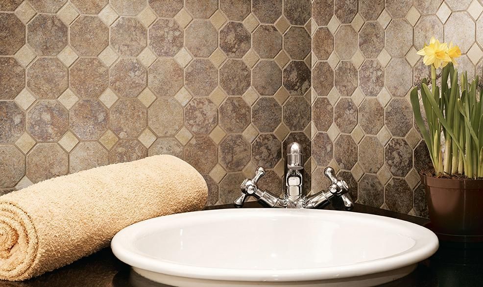 Bathroom sink with white basin, chrome faucet, rolled beige towels, patterned tile, and a potted yellow flower