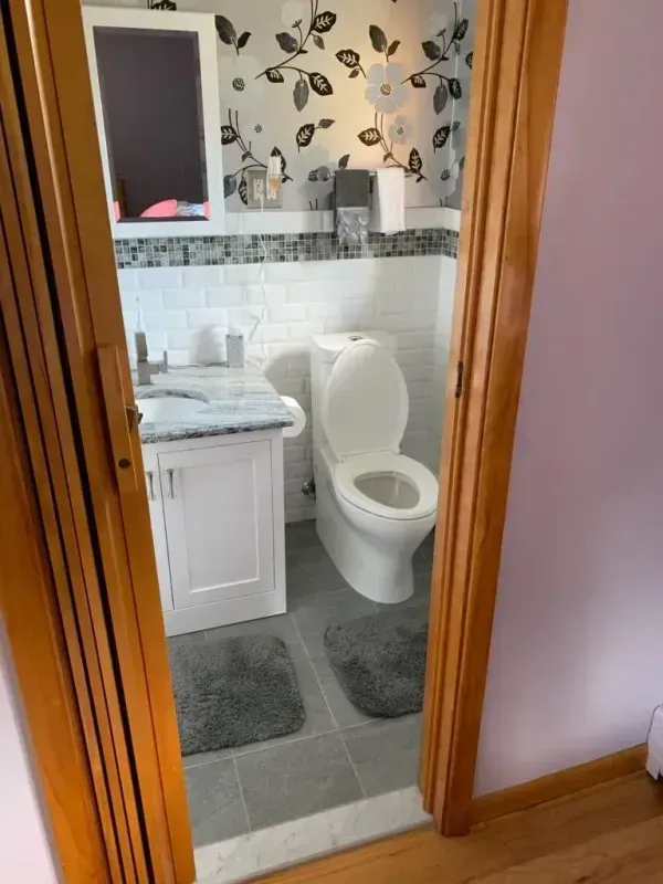 Bathroom with white toilet, sink cabinet, gray rugs, and floral wallpaper, viewed through a wooden doorway