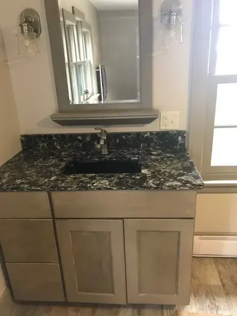 Bathroom vanity with black granite countertop, dark sink, mirror, and light wood cabinet doors beside a door.