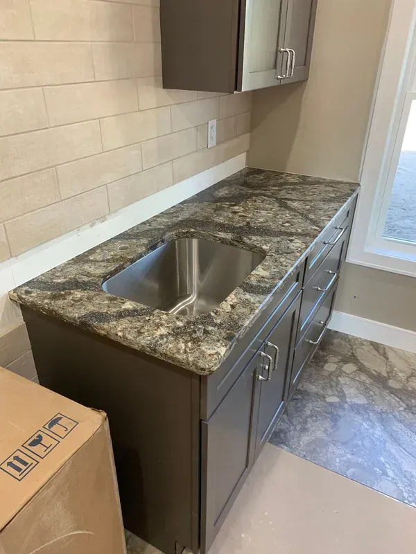 Compact kitchen with granite countertop, stainless sink, gray cabinets, and tiled backsplash near a window.