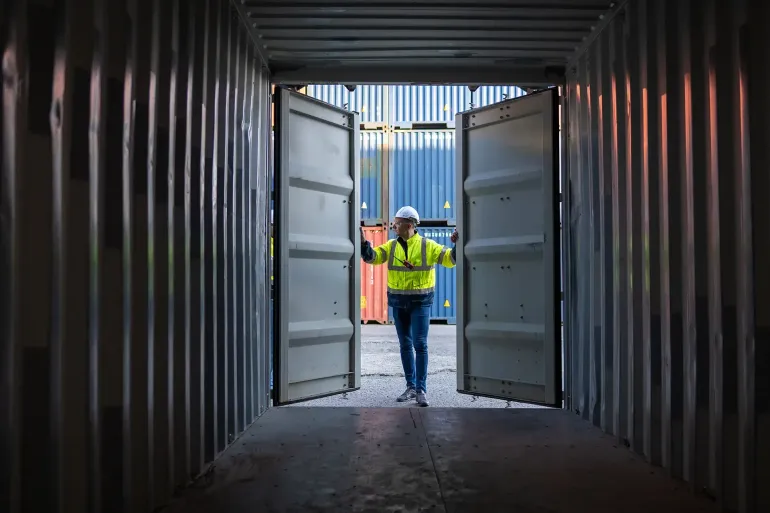 Worker in a high-visibility vest and hard hat walks through an open shipping container.