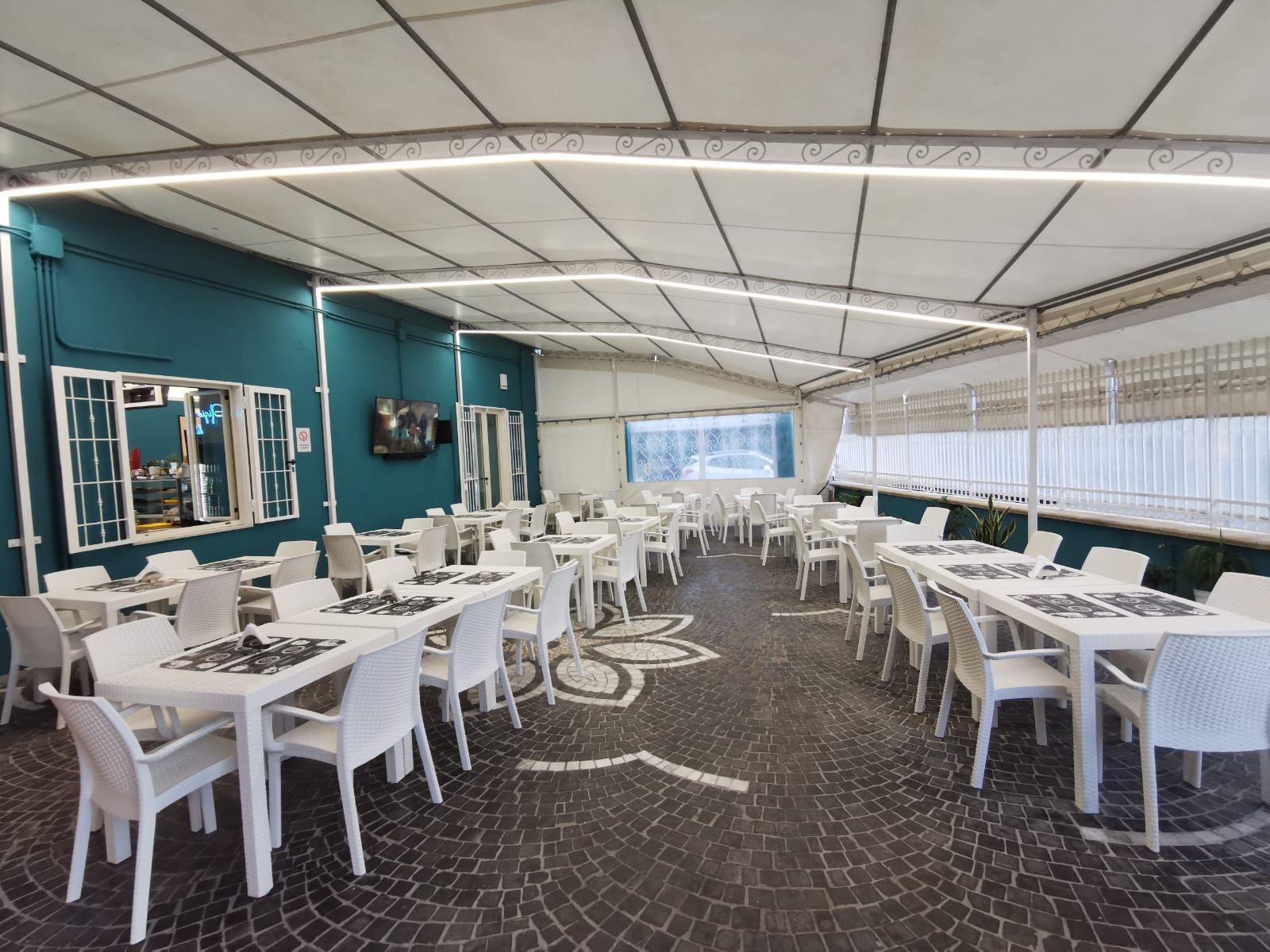 Patio del ristorante con tavoli e sedie bianchi, parete verde acqua e pavimento decorato sotto una tettoia.