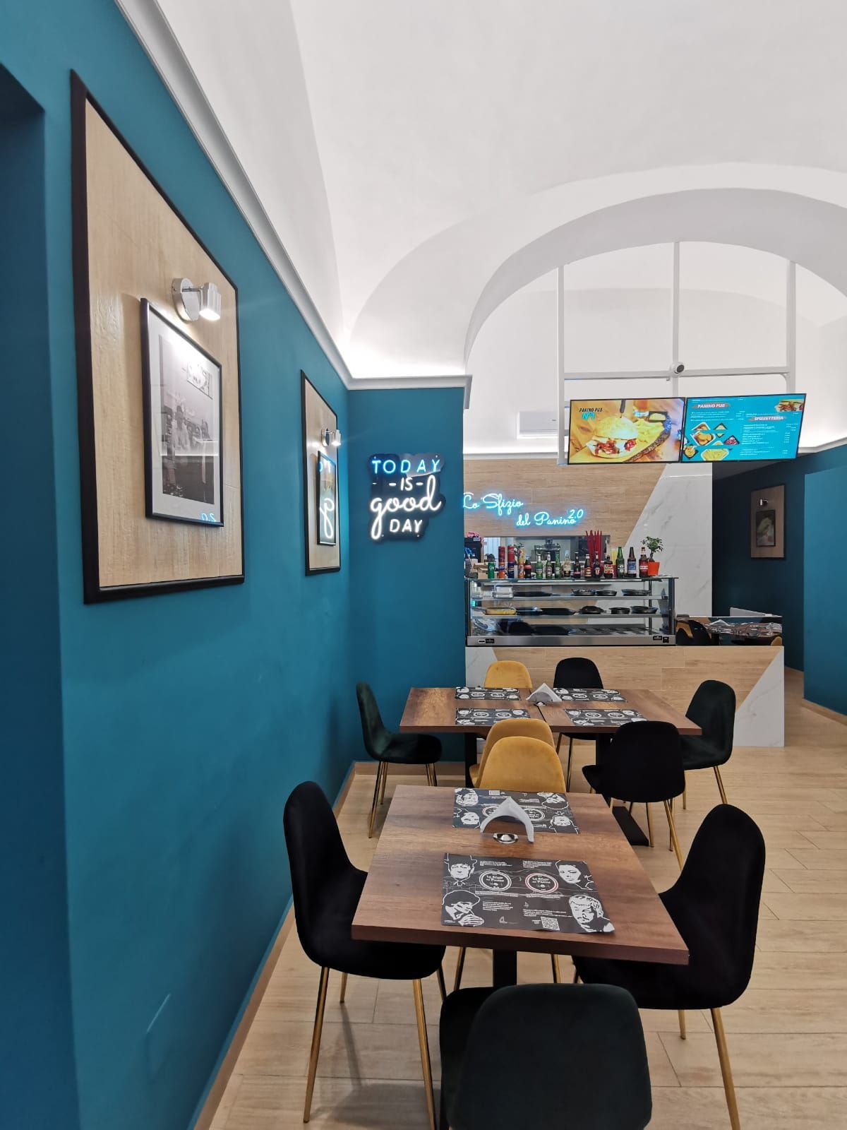 Vista interna di un bar con pareti color verde acqua, tavoli in legno, sedie nere e un'insegna al neon che recita 