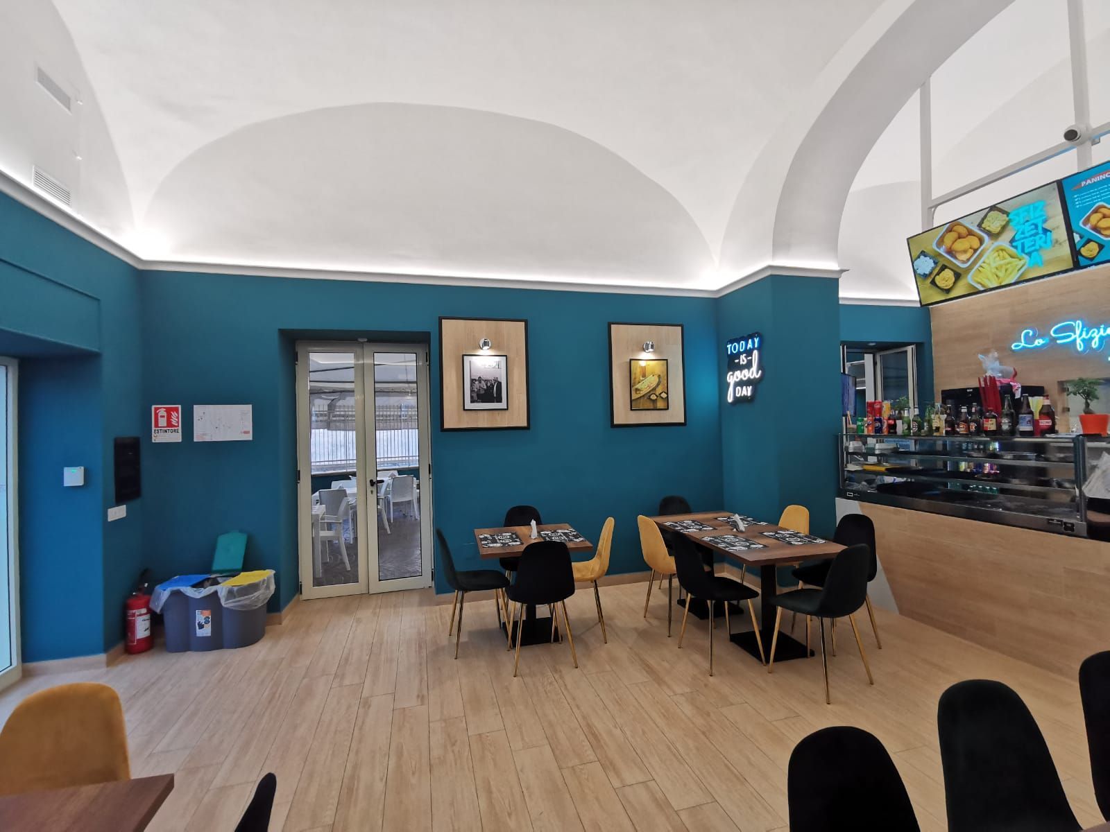 Interni del ristorante: pareti color verde acqua, soffitto bianco a volta, pavimento in legno, tavoli con sedie nere e gialle.
