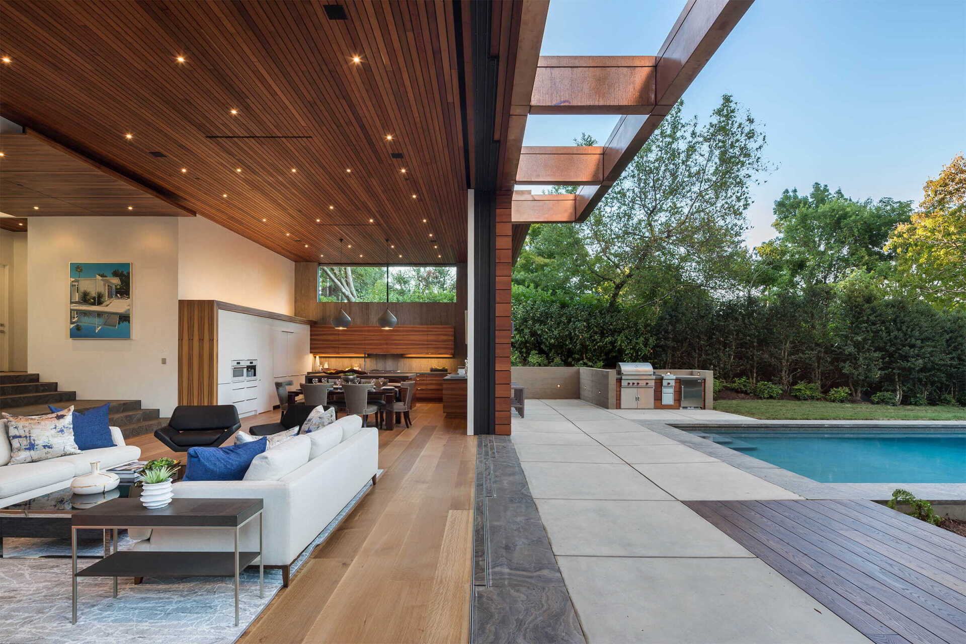 Modern patio with white seating, wood ceiling, and a swimming pool beside greenery