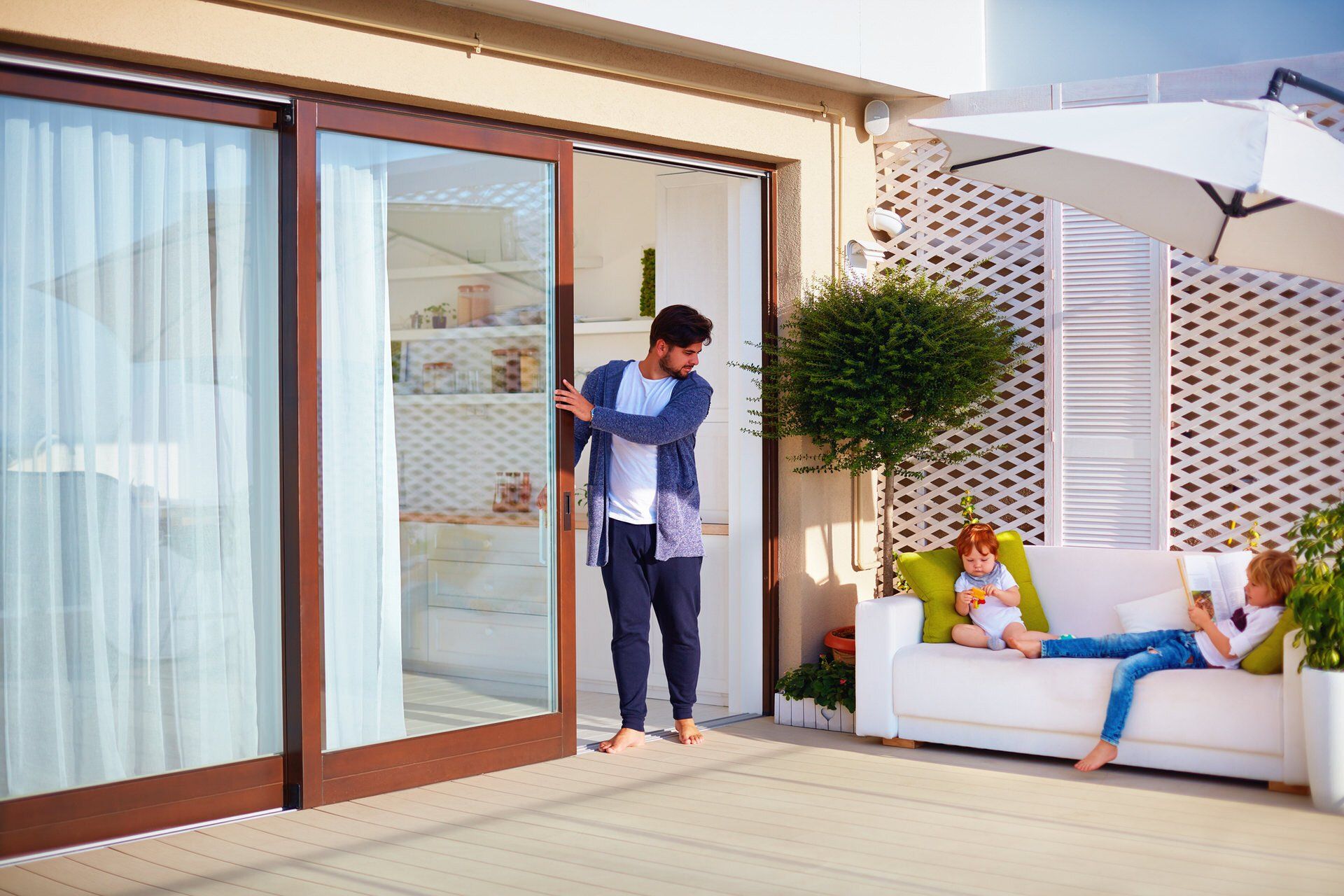 Man on a patio opening a sliding glass door beside a woman lounging on an outdoor sofa