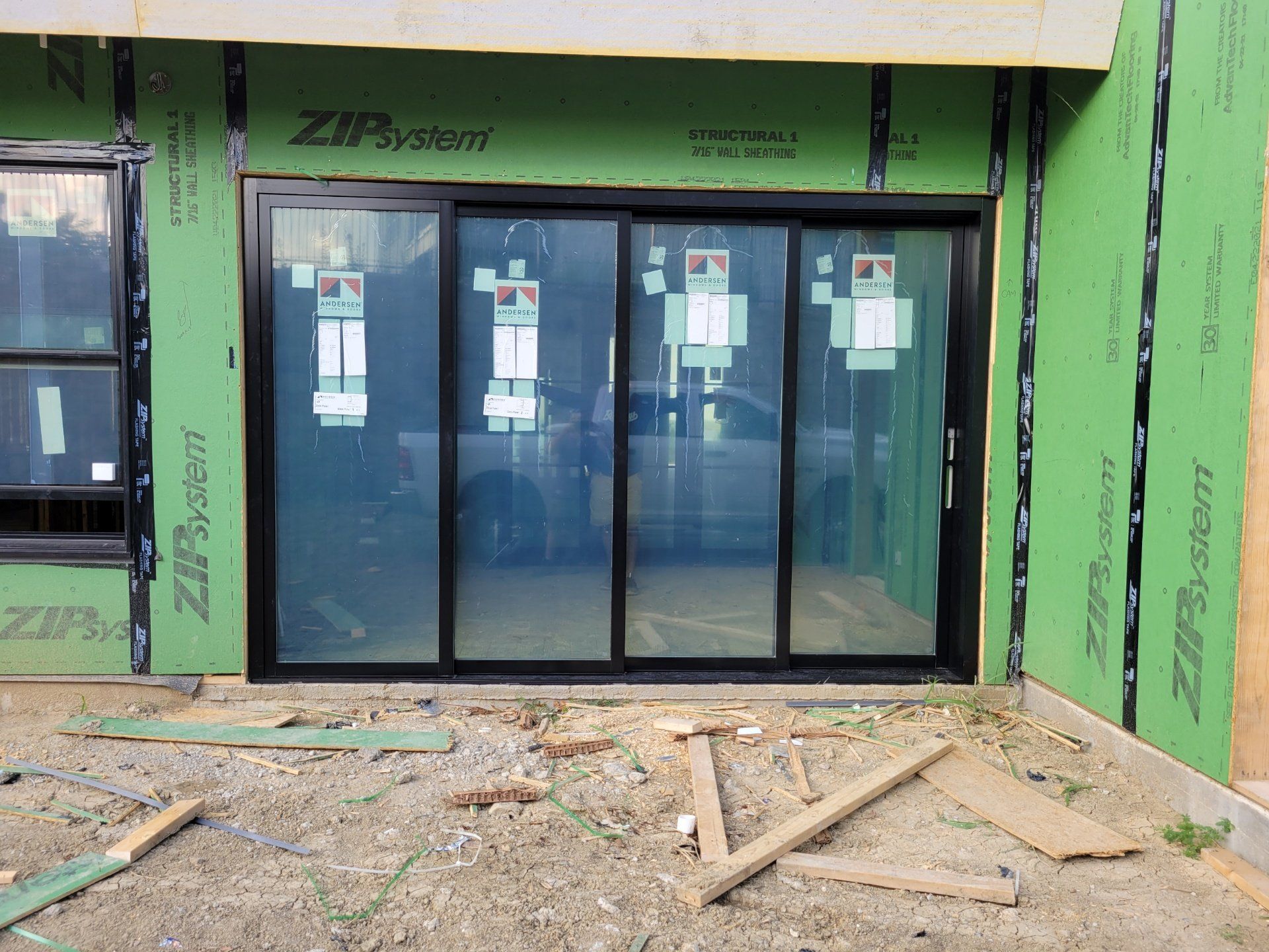 Front entrance with glass sliding doors on a green-wrapped building under construction, muddy ground in front