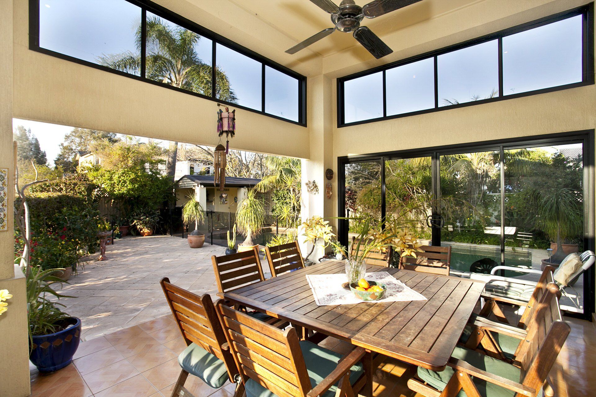 Covered patio with wooden dining table, chairs, ceiling fan, and open view of a sunny garden.