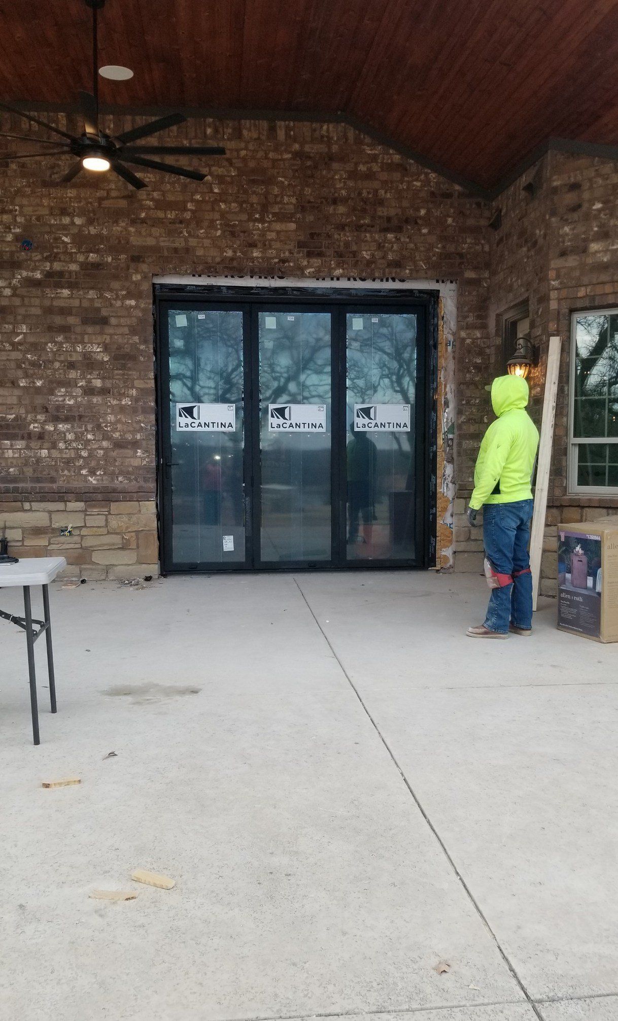 Covered patio with stone walls, concrete floor, ceiling fan, and a person in a neon jacket near glass doors.