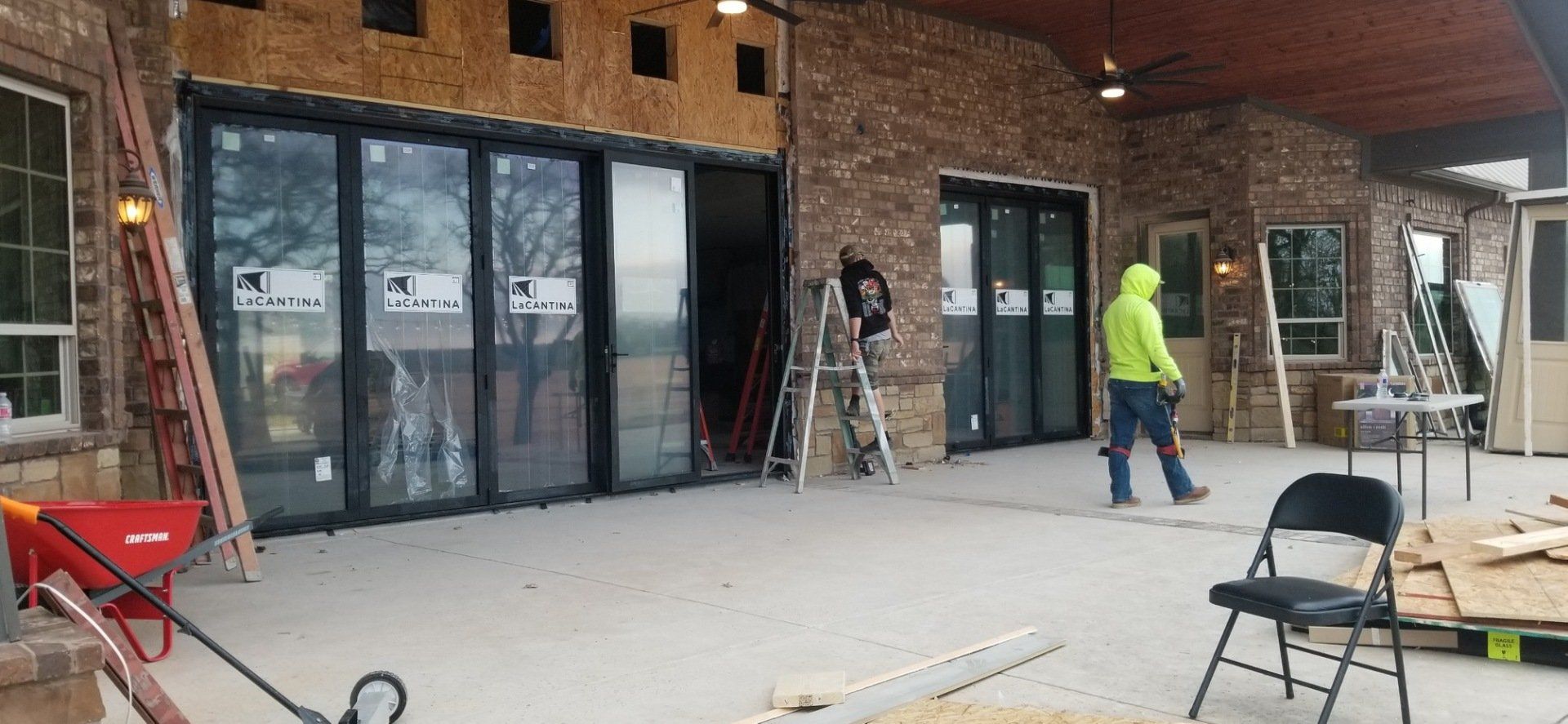 Workers installing glass doors on a brick building porch, with tools and ladders nearby.