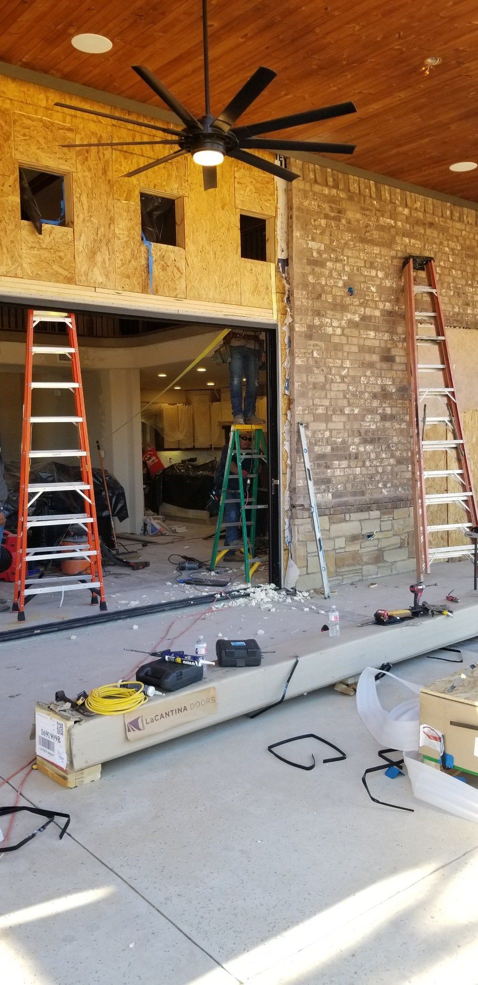 Renovation site with ladders, tools, exposed stone wall, and a black ceiling fan on a wood ceiling