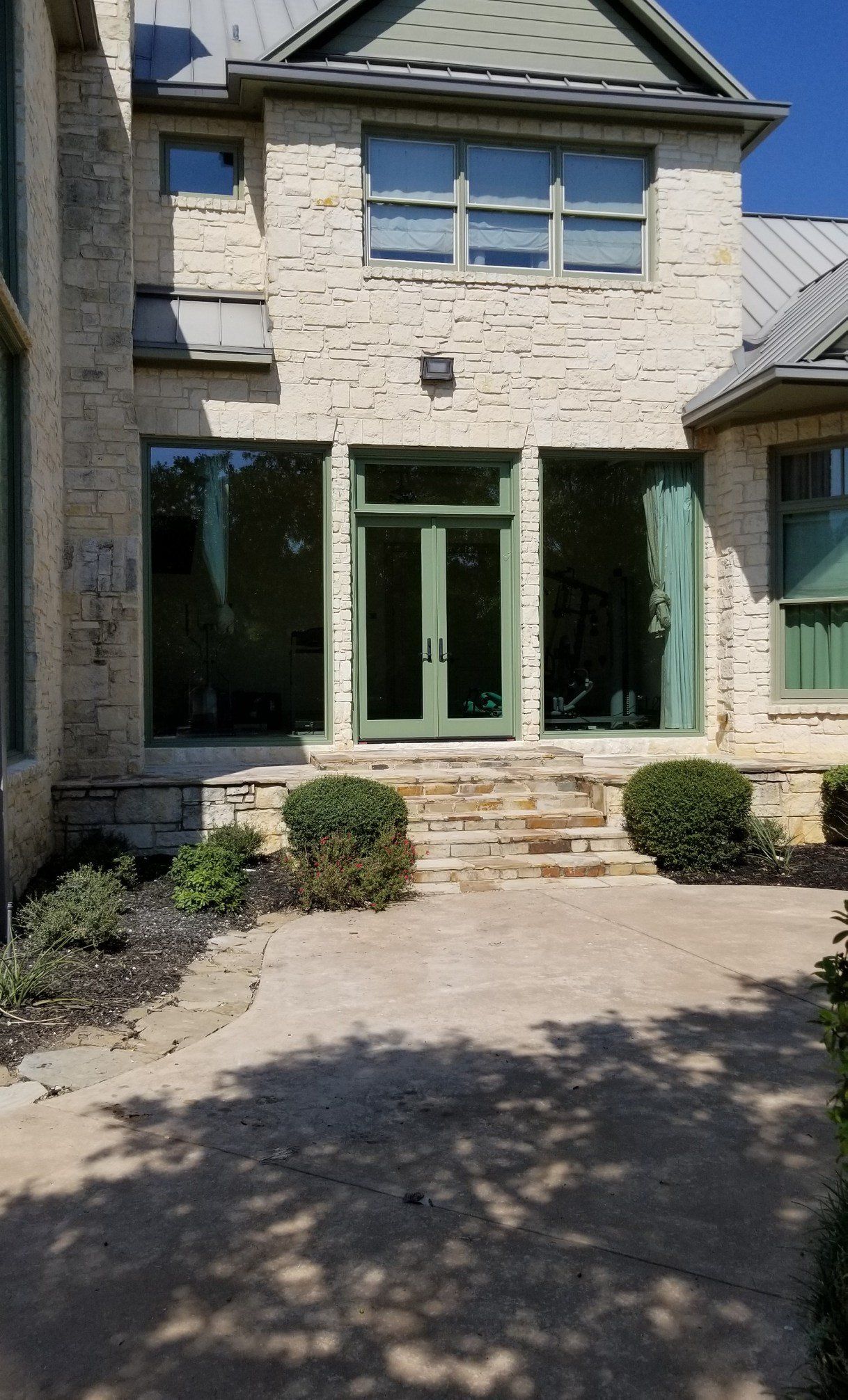 Stone townhouse front with green doors, windows, and a paved driveway under bright sunlight