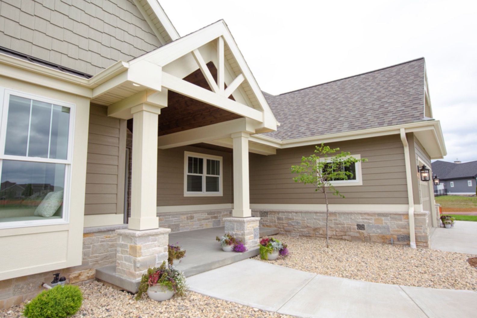 Front entrance of a tan house with white trim, covered porch, stone columns, and a concrete walkway