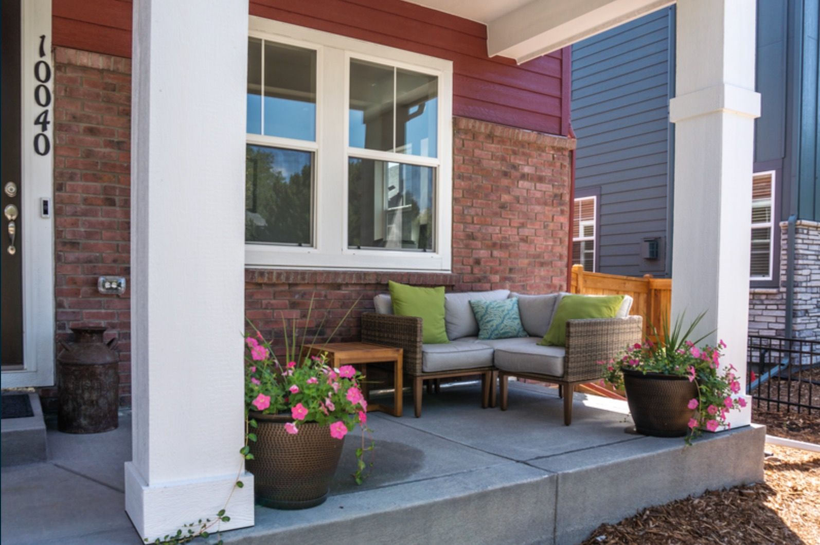 Front porch with wicker chairs, green cushions, potted flowers, and a blue-and-brick exterior.