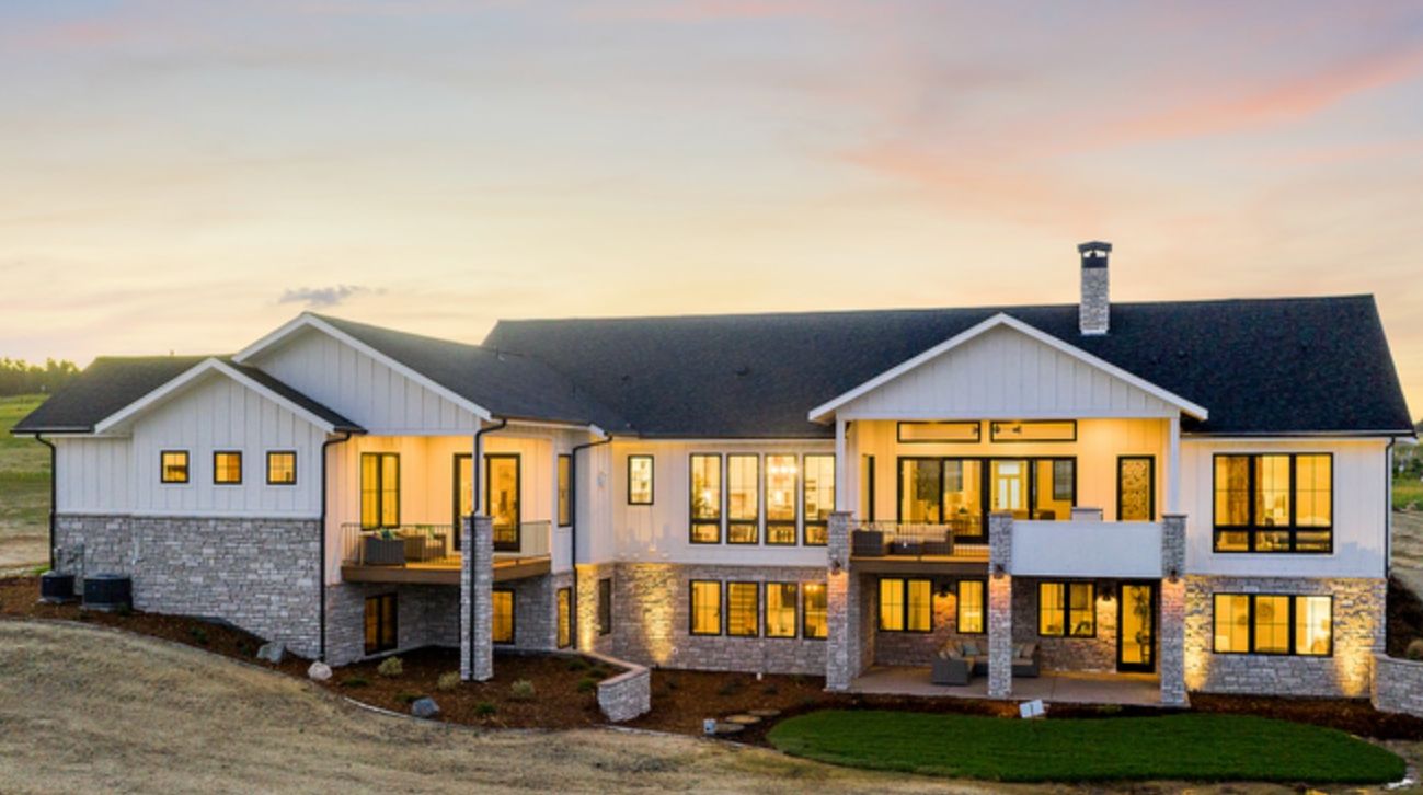 Large two-story beige house with lit windows at dusk and a landscaped front yard