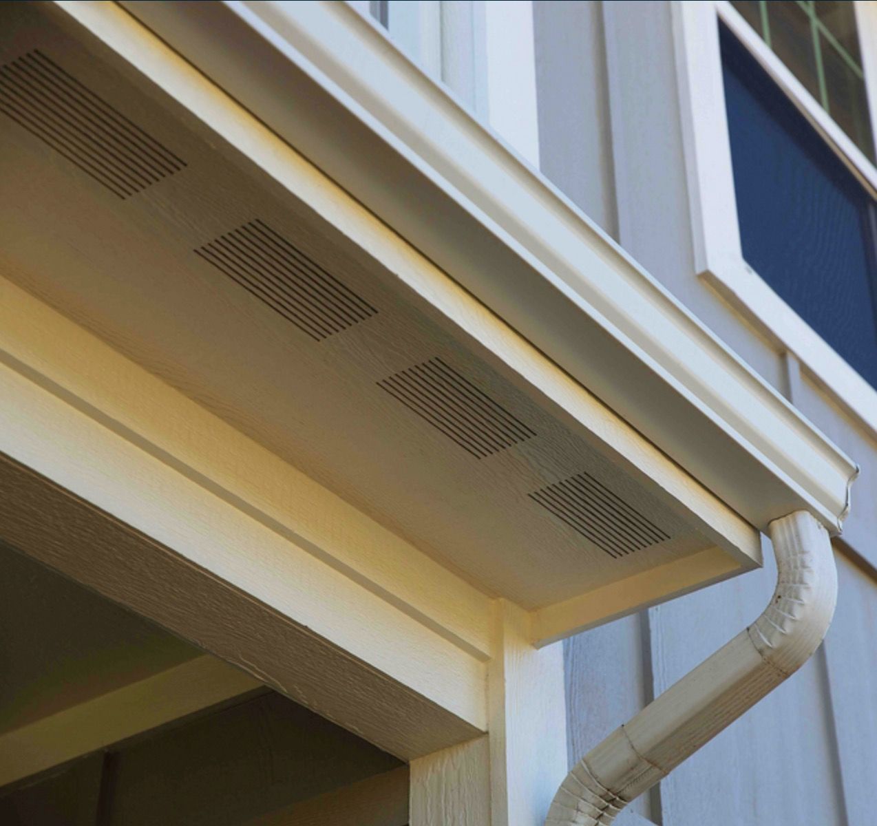 Close-up of beige house eaves with ventilation grilles and a white downspout beside a window