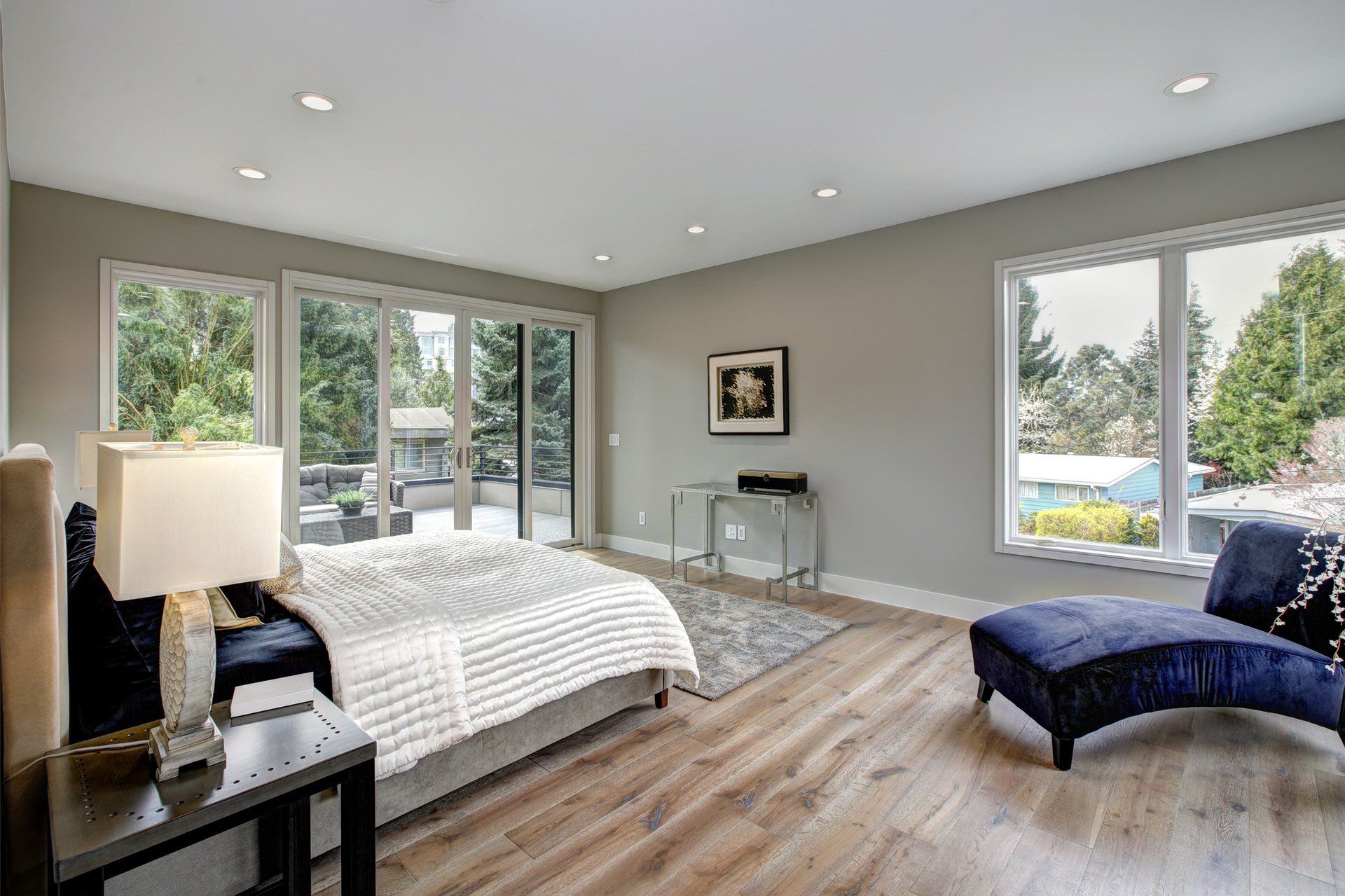 Bright bedroom with large windows, wood floors, a bed, lamp, and blue chaise lounge
