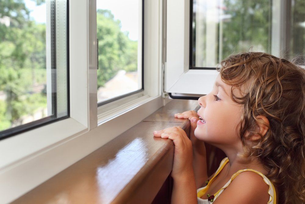 Child looking out an open window, resting hands on the sill in a bright room