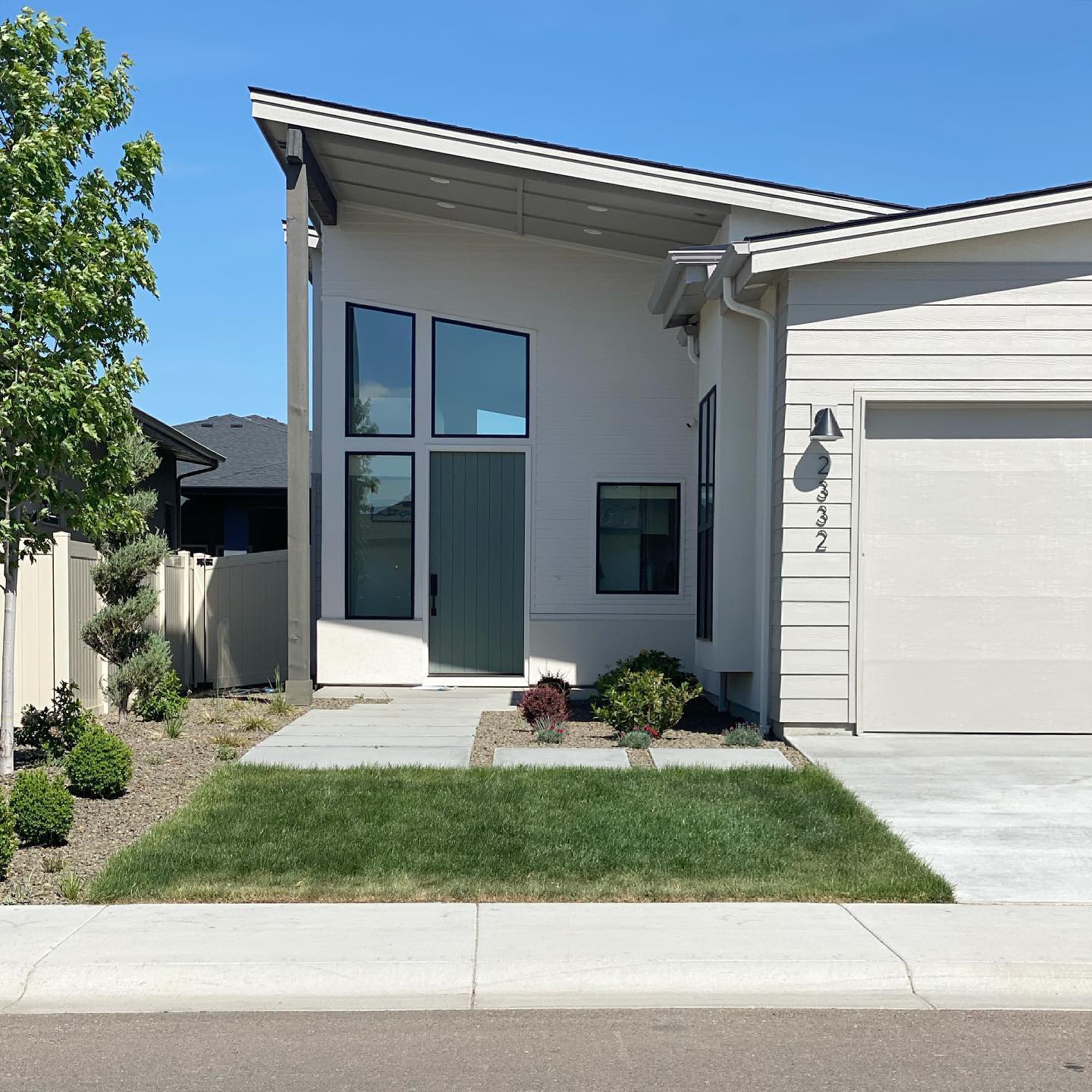 Modern home entry with tall windows, green front door, small porch, lawn, and attached garage under a blue sky