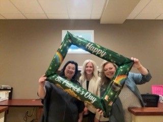 Three people smiling and holding a green “Happy Teacher’s Day” frame indoors