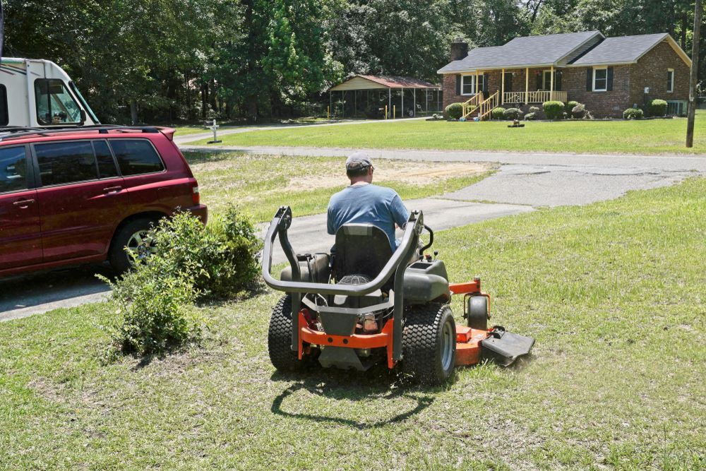 A Man is Riding a Lawn Mower in Front of a House — Oaks Quality Turf In Coolangatta, NSW