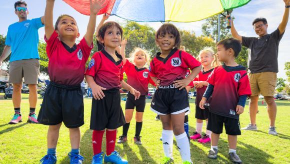 Children in colorful uniforms play a soccer game on an outdoor field.