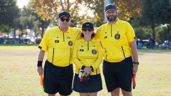 Three soccer referees in matching yellow shirts and black shorts stand on a field, smiling and holding a ball and flags.