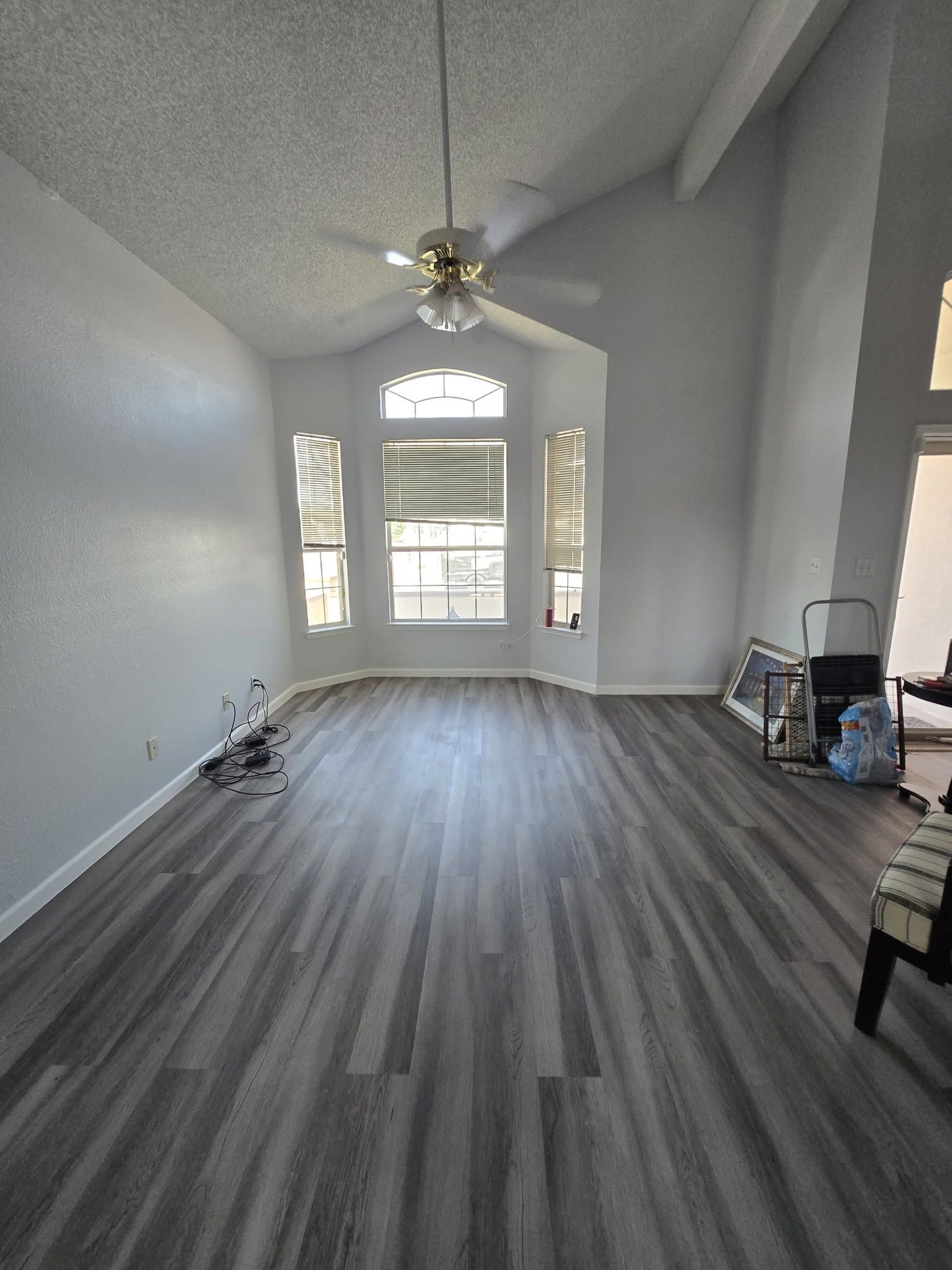 Empty living room with gray wood floors, vaulted ceiling, ceiling fan, and large front windows.