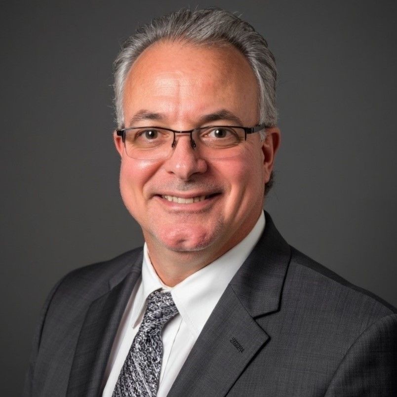 Man in suit and glasses smiles at the camera against a gray backdrop.