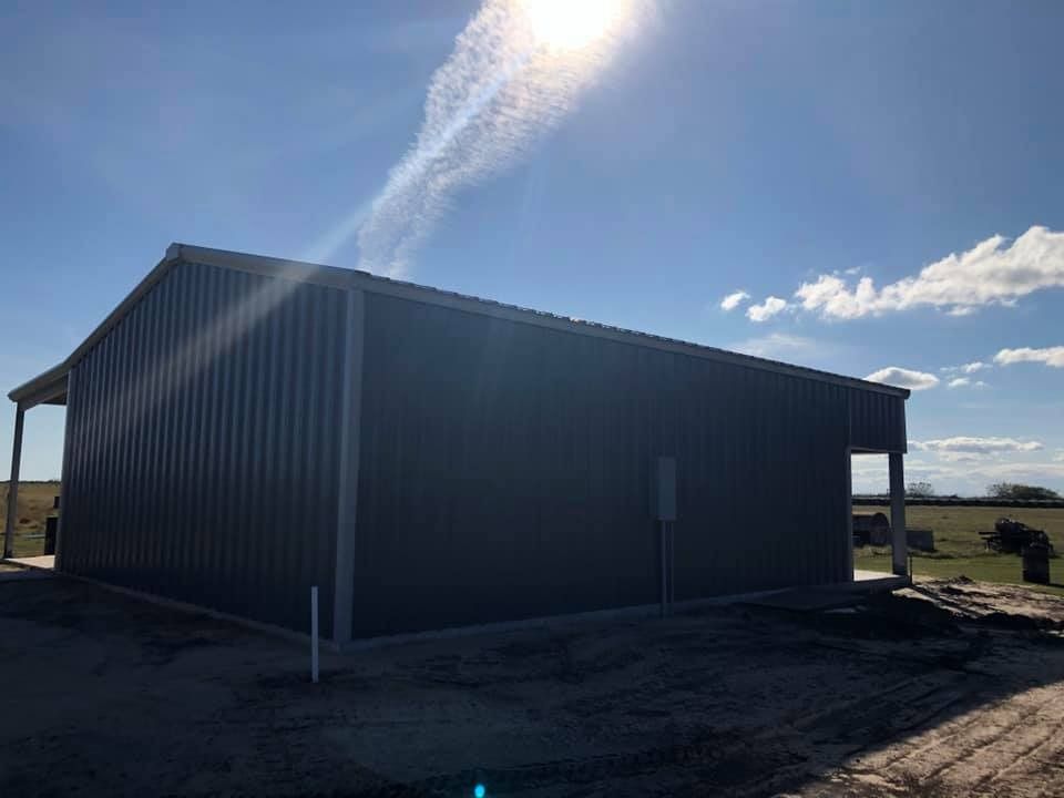 A modern dark gray metal building stands in a grassy rural field under a bright, sunny sky.