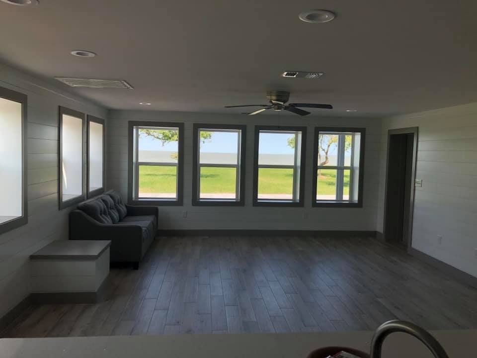 A bright, minimalist living room with gray tile flooring, a dark sofa, and a row of windows overlooking a grassy field.