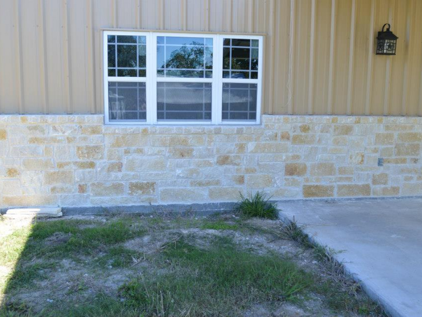 A tan metal-sided building wall with a stone veneer base and a grid-pane window above a small grassy patch and concrete.