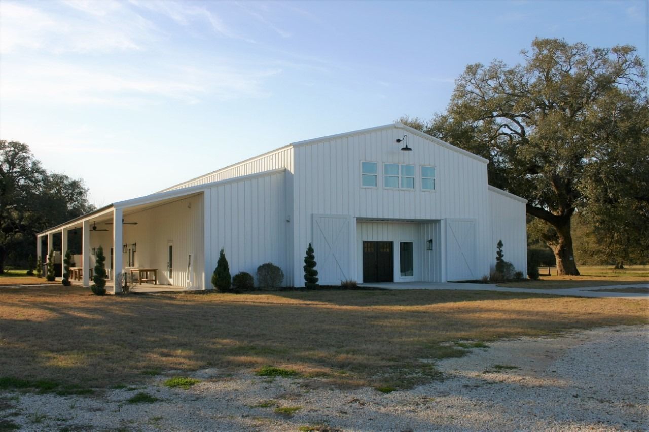 A white, modern pole barn building with a covered side patio sits on a grassy lot with large trees in the background.