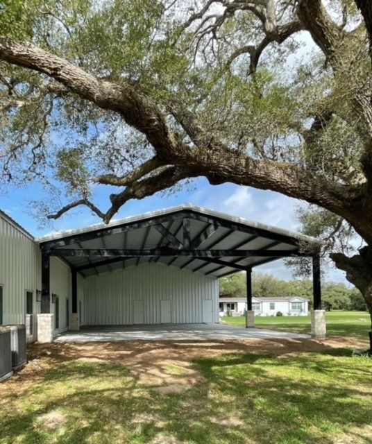 A white metal carport extension attached to a building, partially shaded by the large, overhanging limbs of an oak tree.