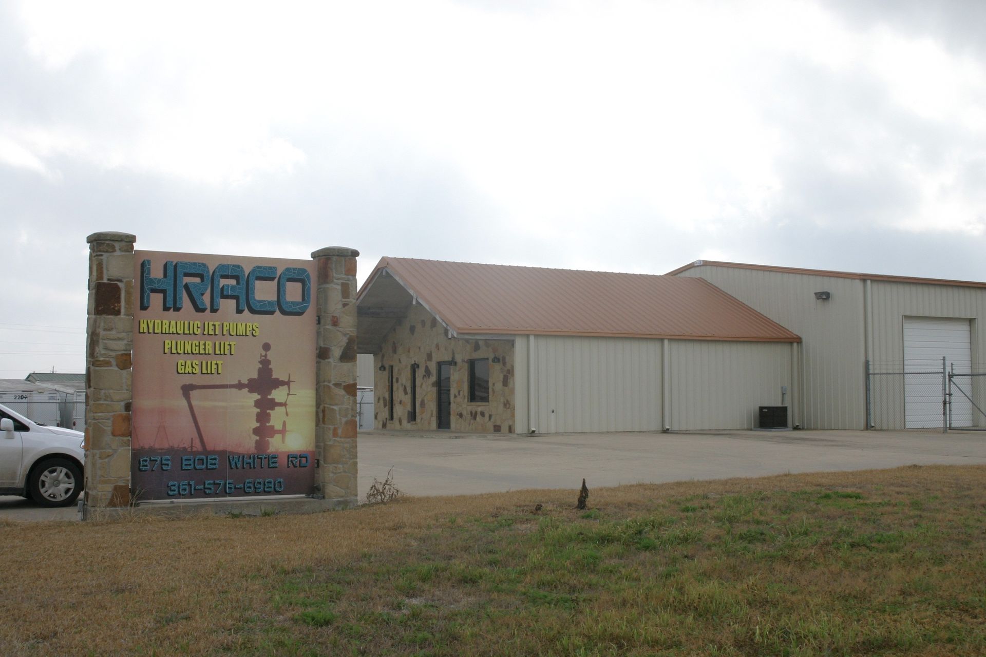 A roadside sign for HRACO in front of a tan industrial office building under a cloudy sky.
