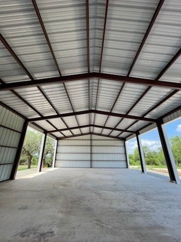 A wide, open-sided metal pole barn with a concrete floor, gabled roof, and dark brown support beams.