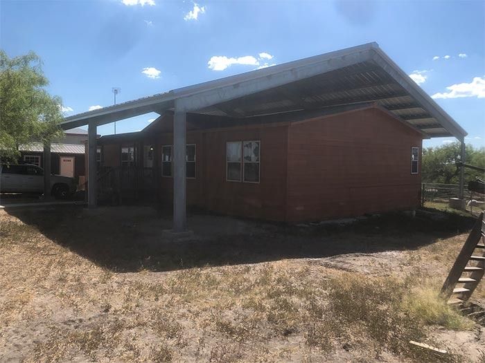 A tan manufactured home with a large metal carport cover over its side and roof, set on a grassy lot under a sunny sky.