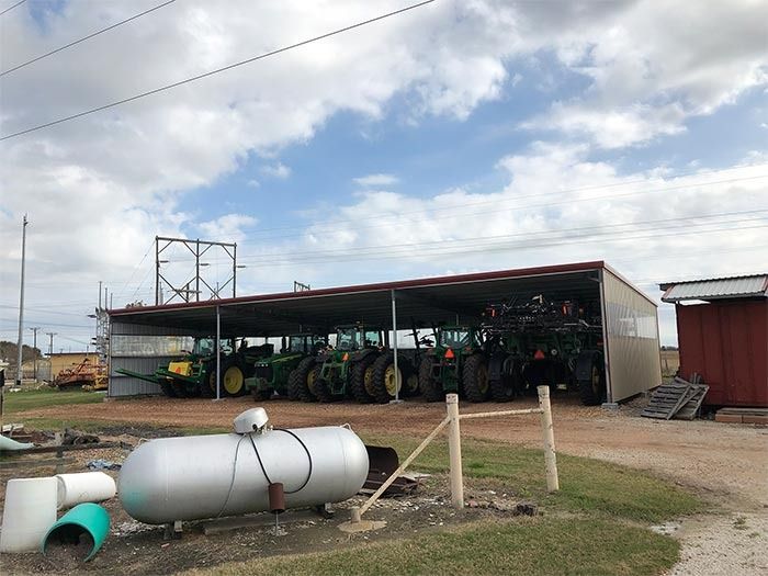 A row of green farm tractors parked inside an open-sided metal shed under a cloudy sky.