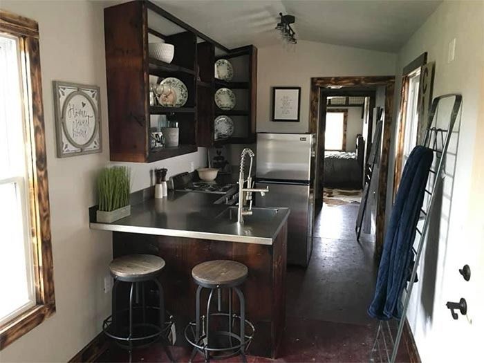 A rustic kitchen featuring dark wood cabinets, stainless steel counters, two bar stools, and an open doorway.