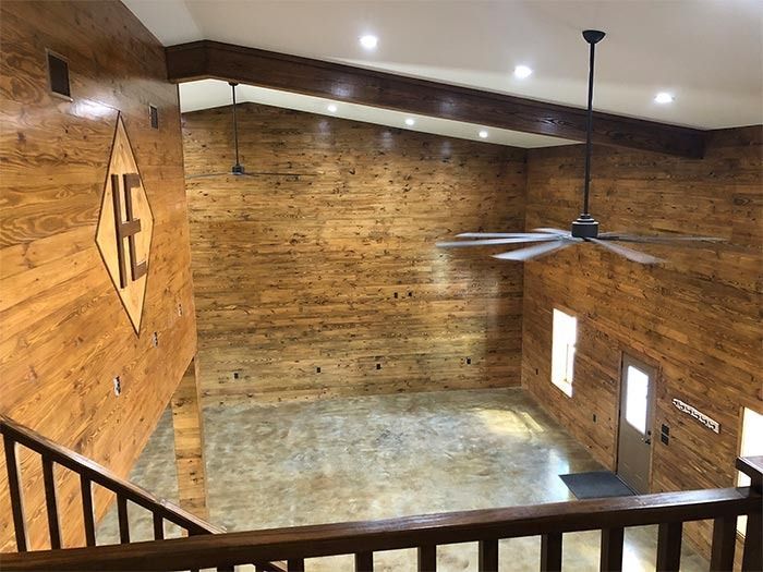 An interior view of a tall room with wood-paneled walls, a sloped ceiling, concrete floors, and a hanging ceiling fan.