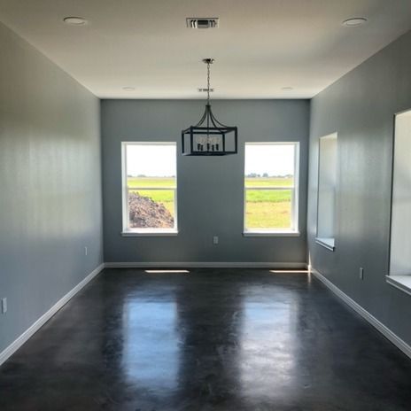 A minimalist room with grey walls, polished dark floors, two windows overlooking a field, and a black metal chandelier.
