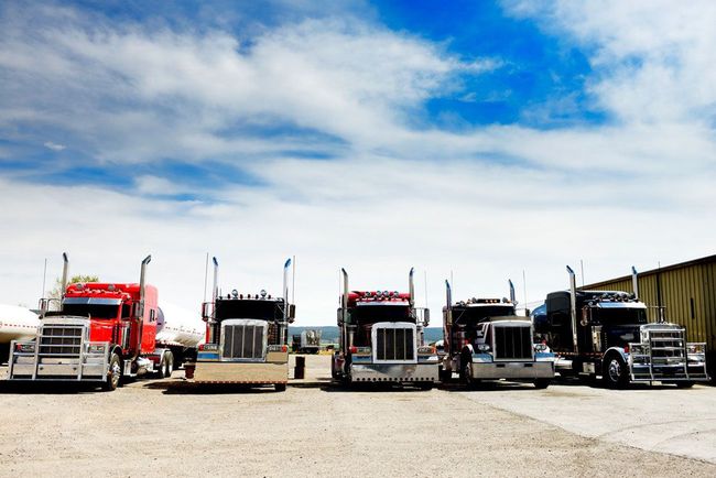 Five semi-trucks parked in a row on a gravel lot under a bright, cloudy sky.