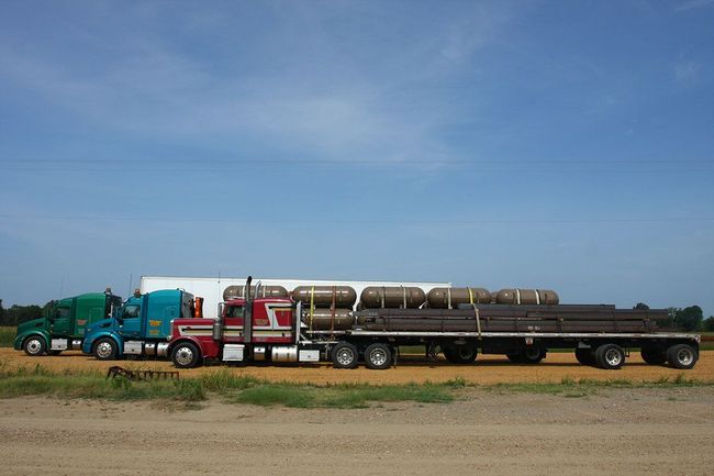 A row of three parked semi-trucks—green, blue, and red—on a dirt lot under a bright blue sky, with cargo on a flatbed.