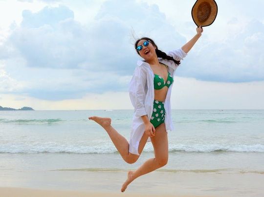 Woman jumps on beach, wearing polka dot bikini, holding hat, smiling.