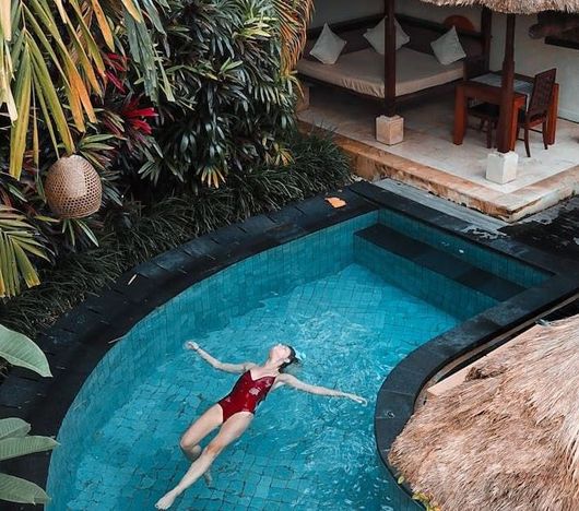 Woman in red swimsuit floats in a blue pool. Tropical setting with thatched roof and outdoor lounge.