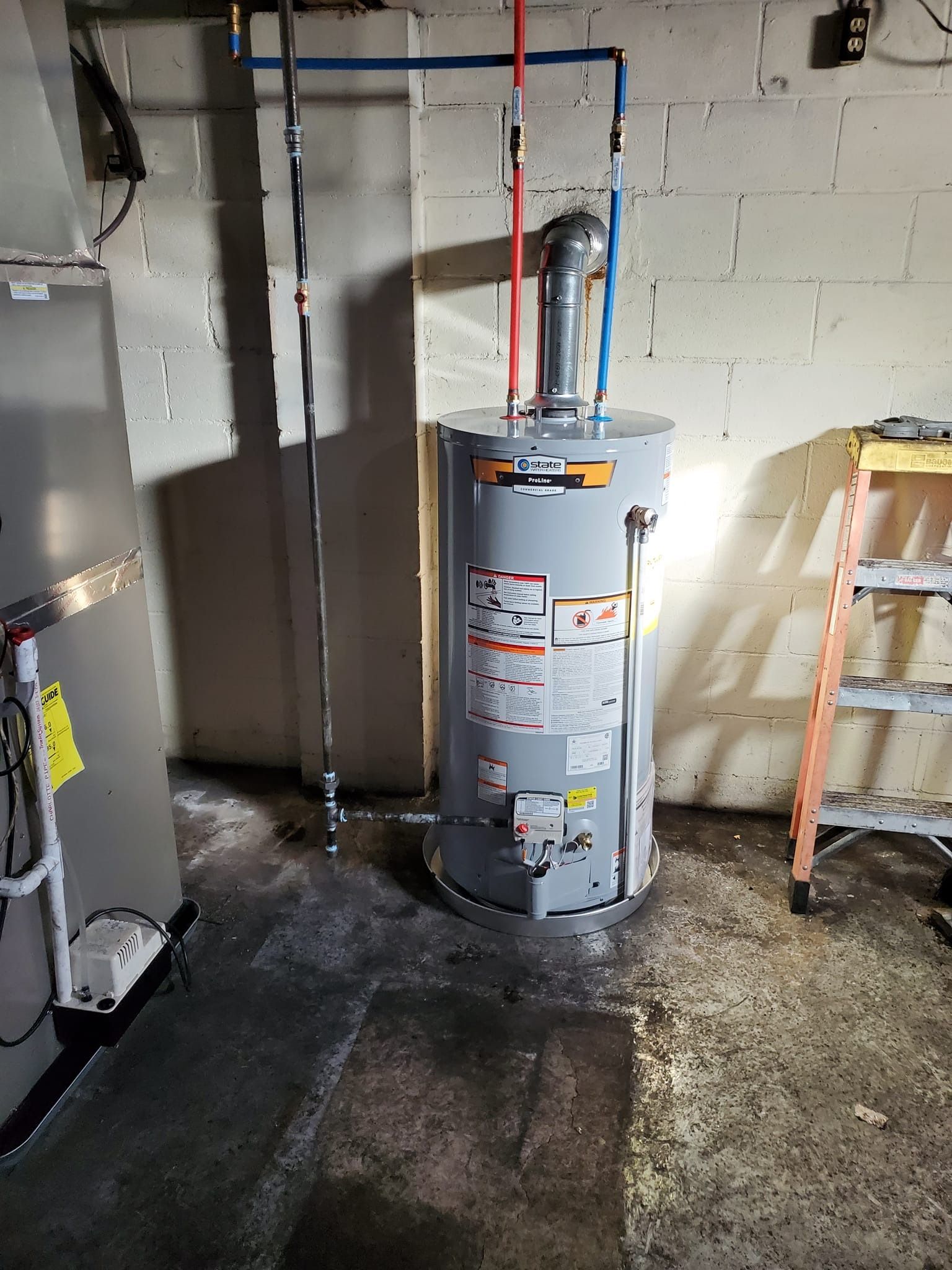 A water heater is sitting in a basement next to a ladder.