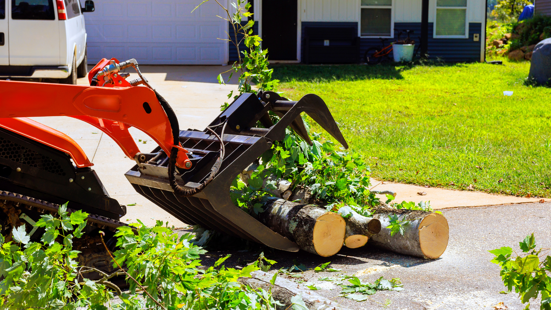 Orange skid steer lifting cut logs and branches on a suburban driveway