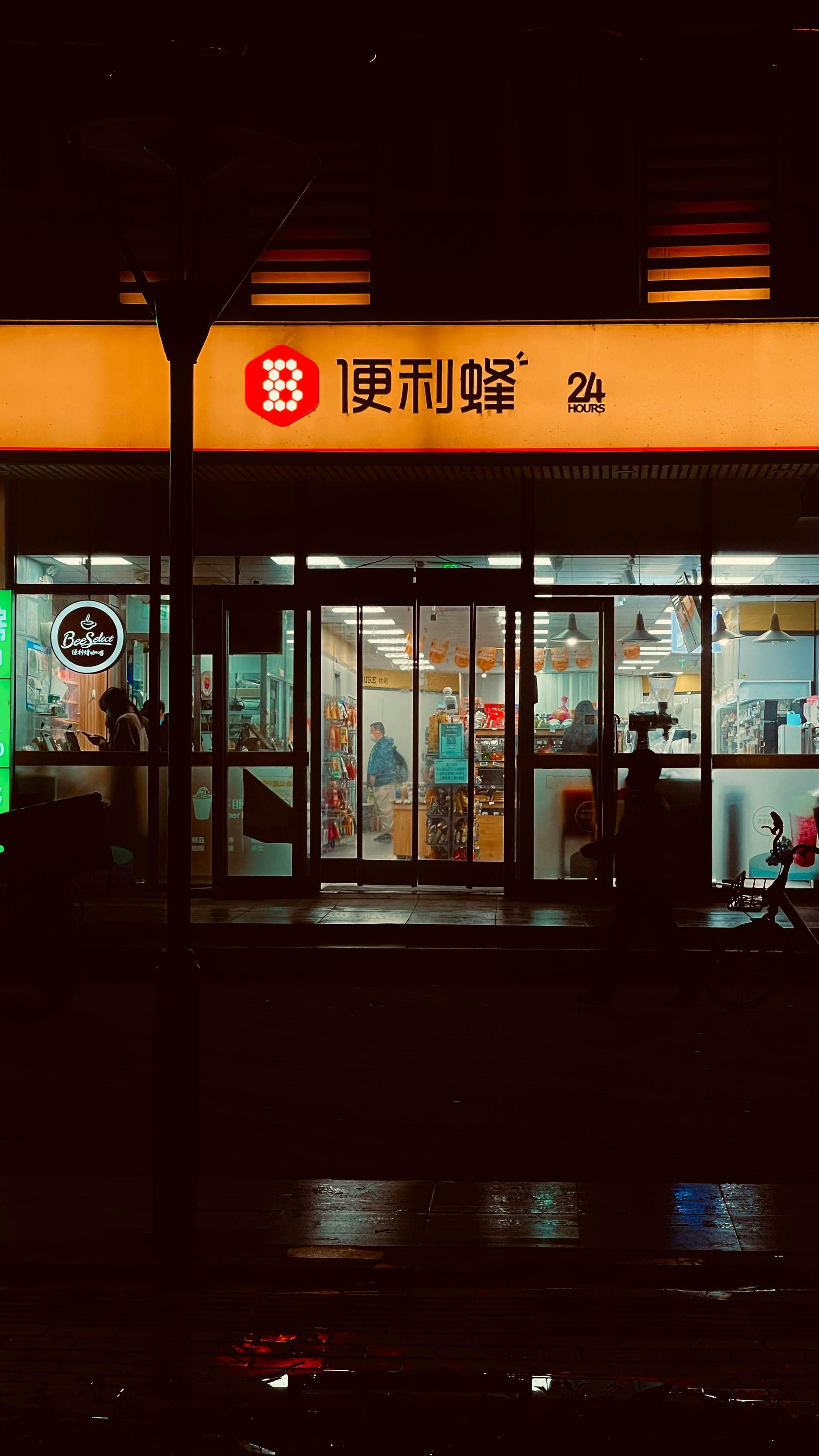 Night view of a brightly lit convenience store with Chinese characters above the entrance. Reflective street below.