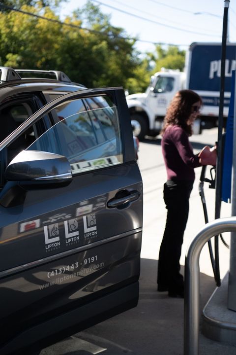 Woman filling gas tank of gray car at a gas station. White truck in the background.