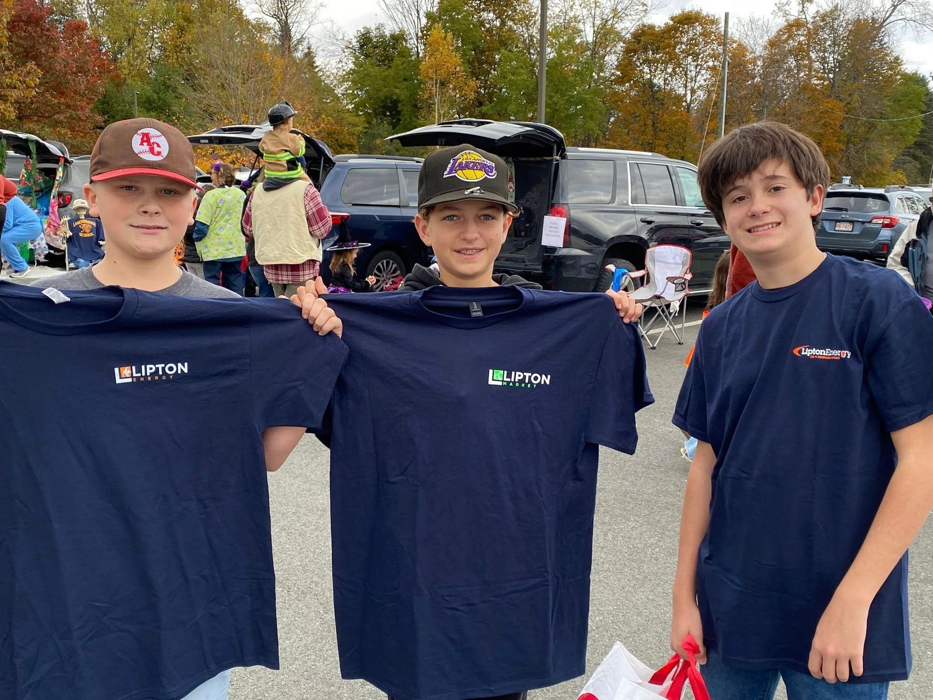 Three boys hold up navy blue T-shirts with a logo, smiling in an outdoor setting.